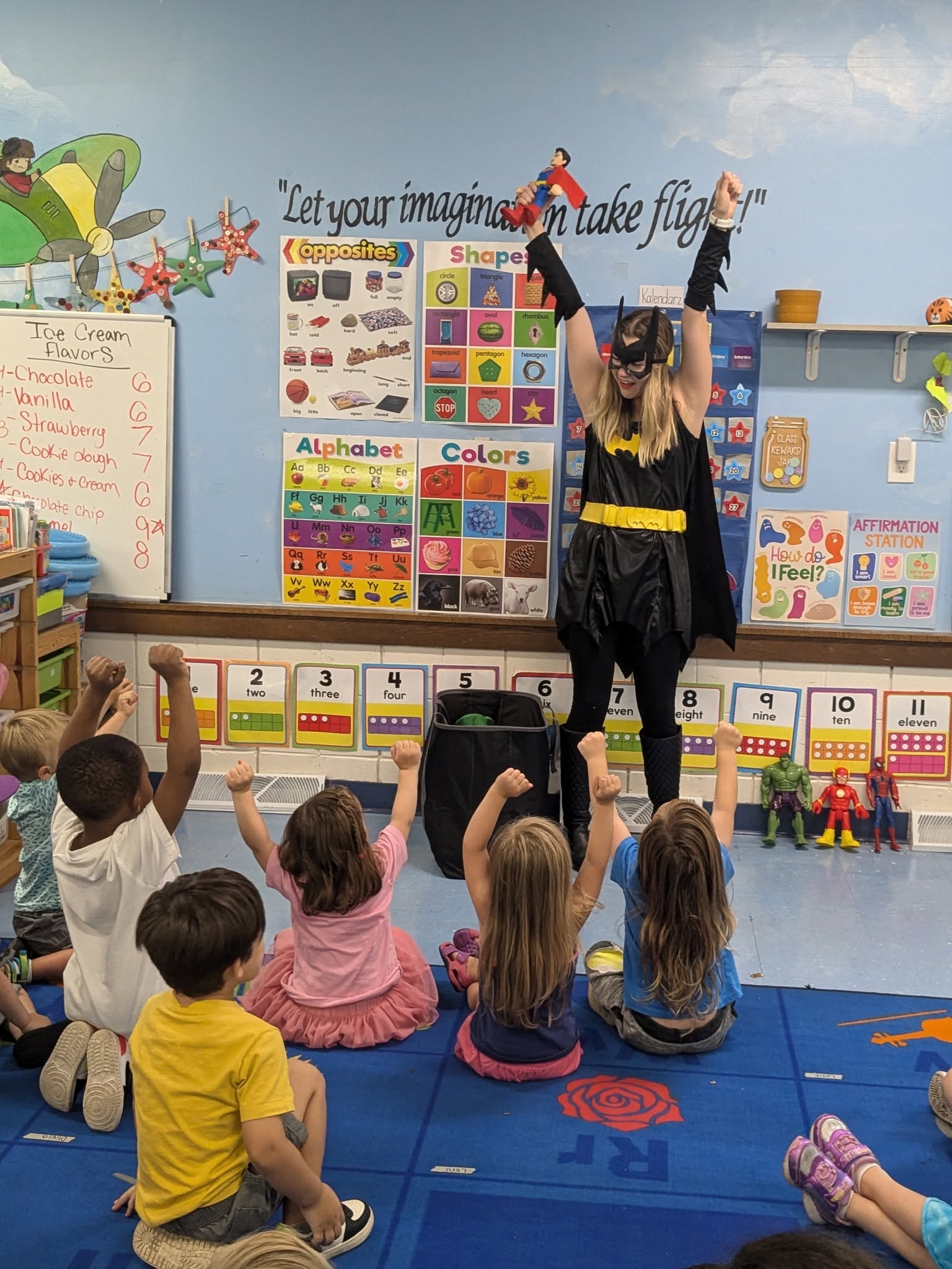 A teacher dressed as a superhero with cheering school-age kids at Creative Kids Academy Maple Grove