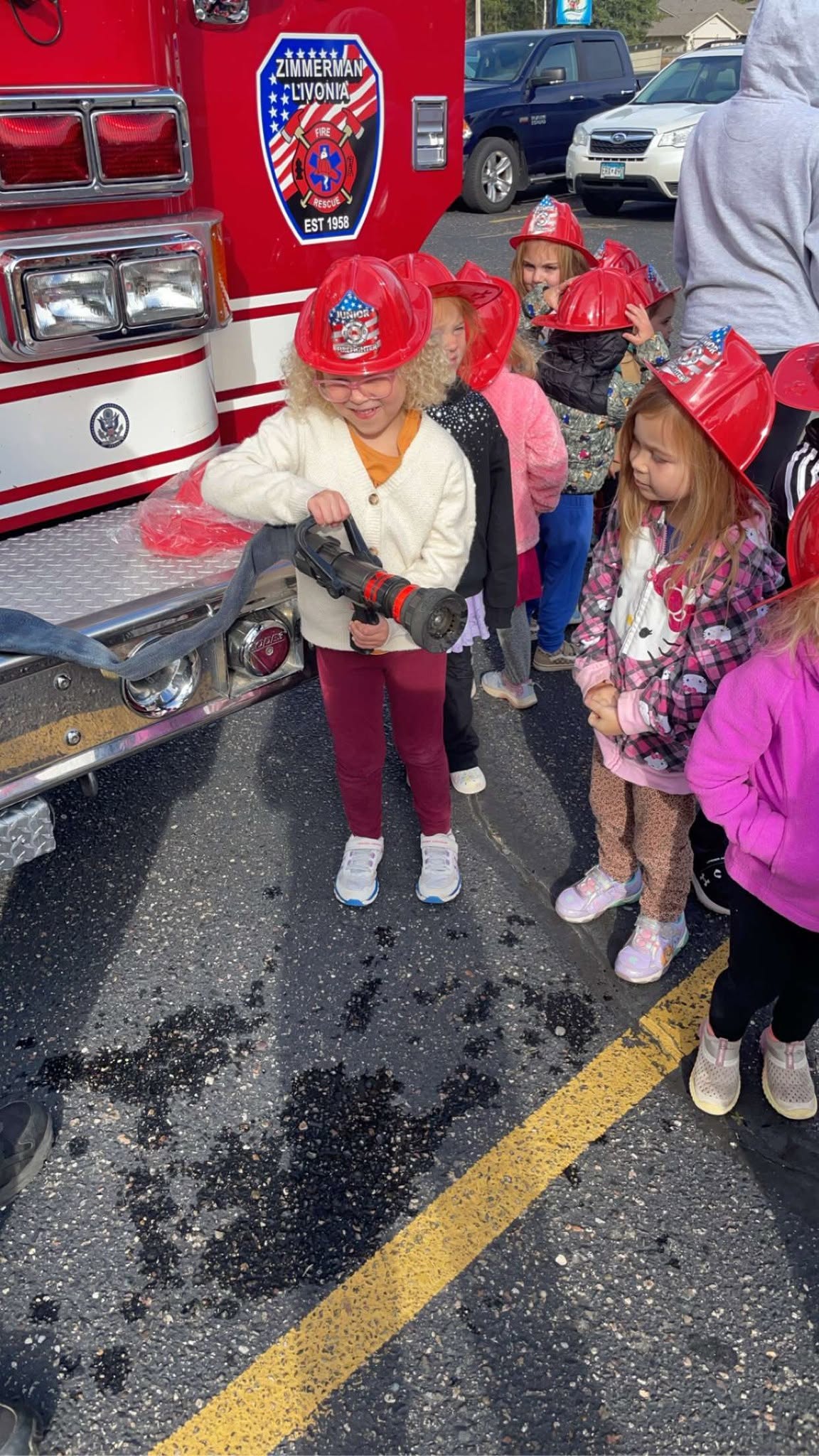 Children in junior firefighter helmets exploring a fire truck outside Creative Kids Academy Zimmerman