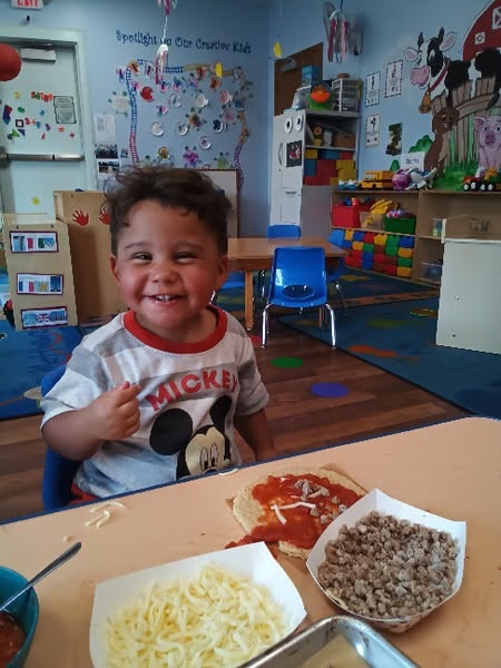 A toddler smiling while making a pizza during a cooking activity at Creative Kids Academy Maplewood
