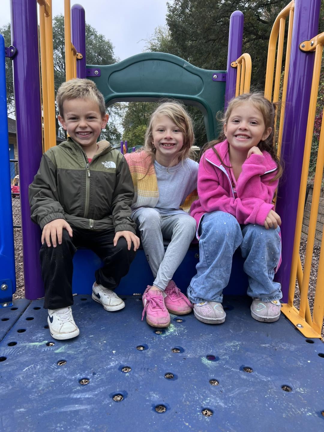 Three smiling children sitting together on outdoor playground equipment at Creative Kids Academy Elk River on 10