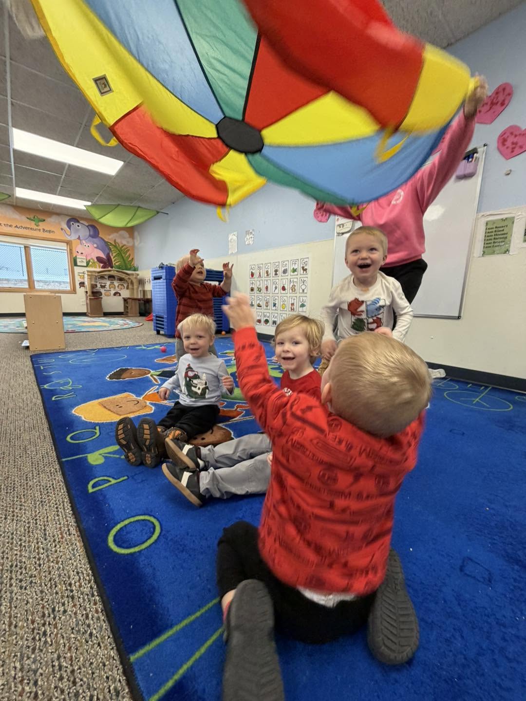 Toddlers reaching up to play with a rainbow parachute during circle time at Creative Kids Academy Elk River on 10