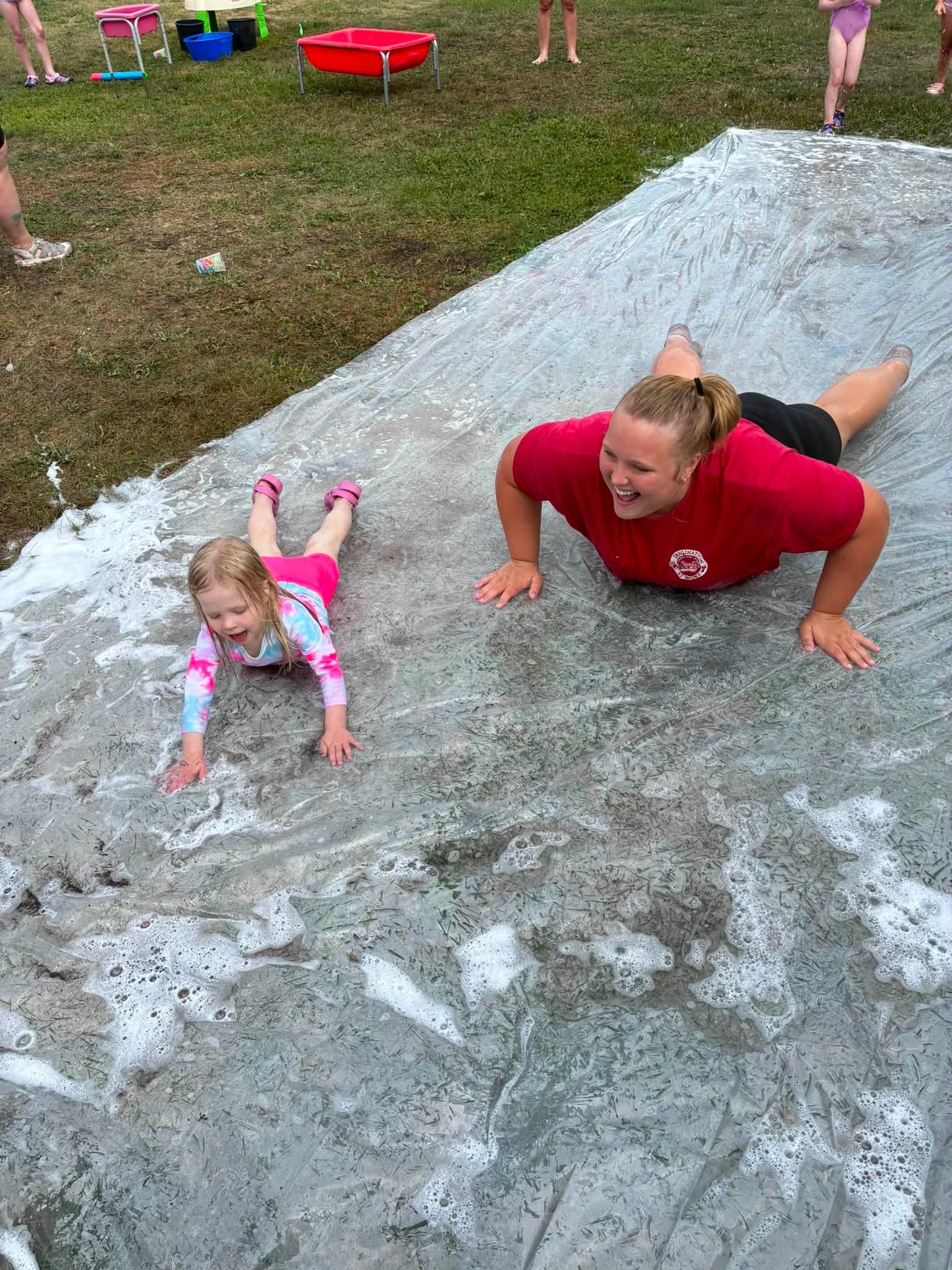 A teacher and preschooler laughing together on a soapy slip-n-slide at Creative Kids Academy Elk River