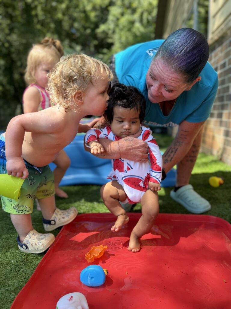 A teacher helping a baby stand in a water table while a toddler leans in to kiss the baby