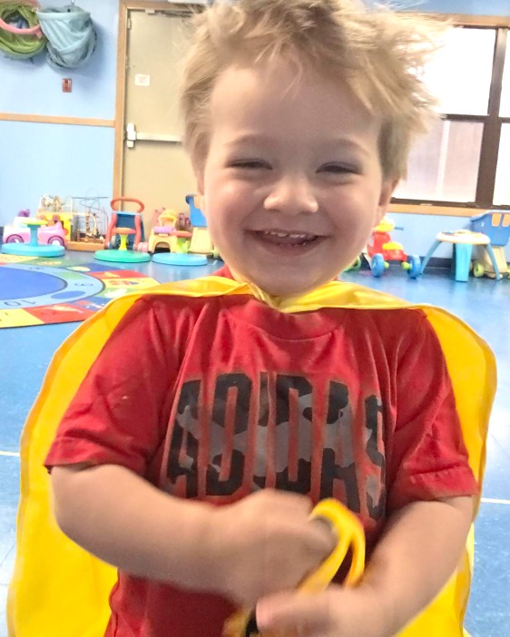 Toddler in a superhero cape smiling in an Emerging Toddler classroom at Creative Kids Academy