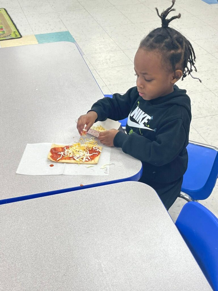 Toddler adding toppings to a mini pizza during a CKA cooking activity