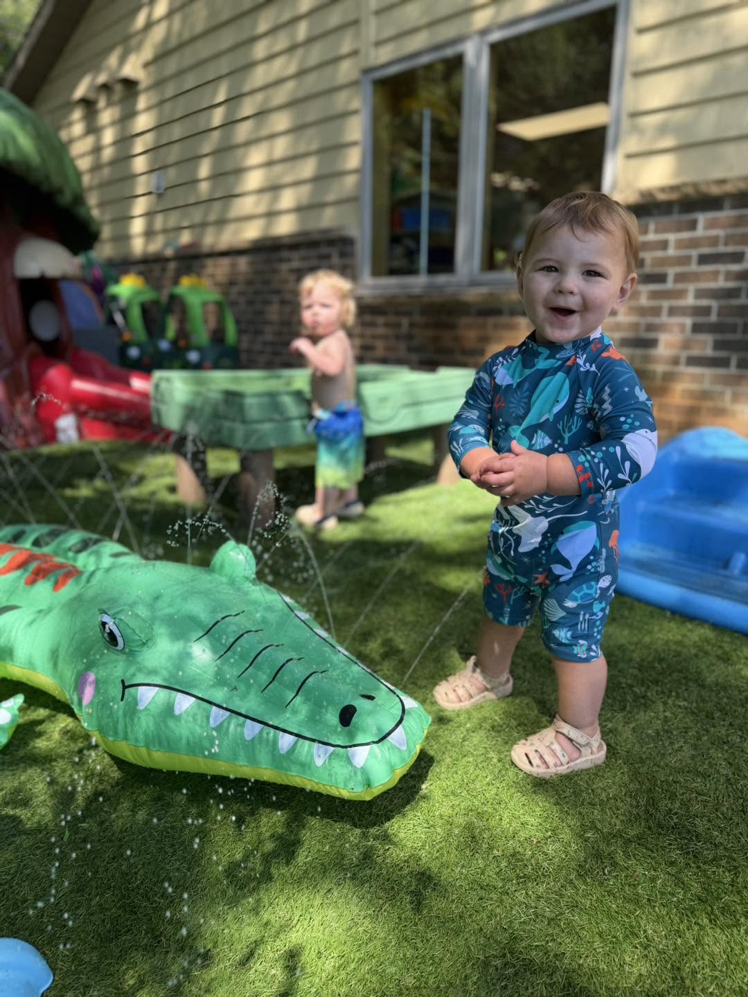 Toddler smiling next to a crocodile sprinkler during outdoor water play at Creative Kids Academy